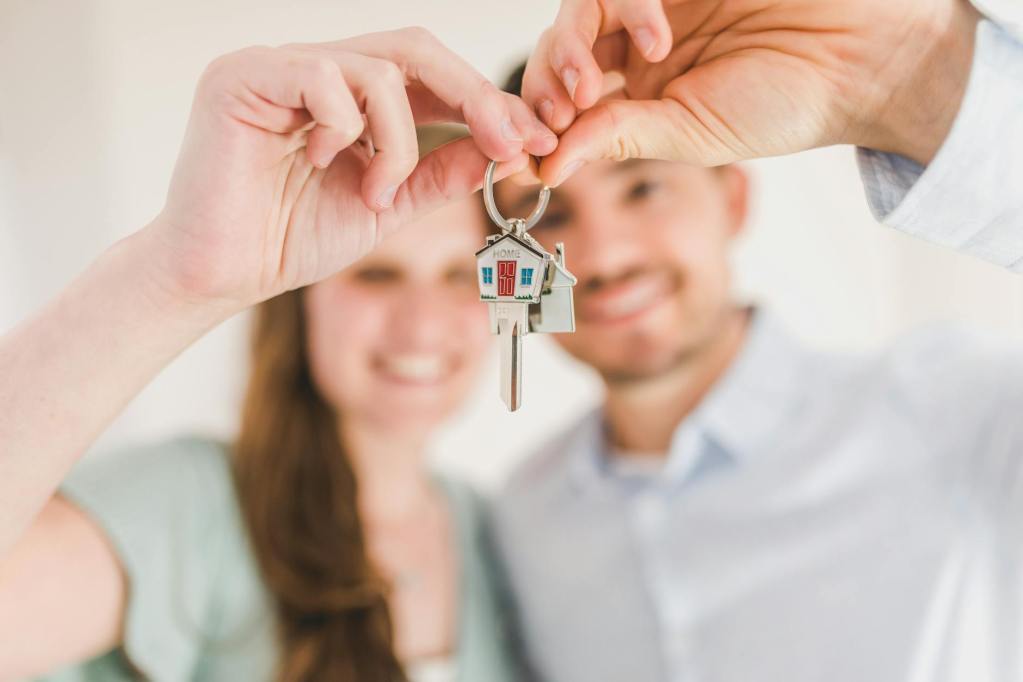 A couple holding a house key with a keychain shaped like a house, smiling in a bright indoor setting.
